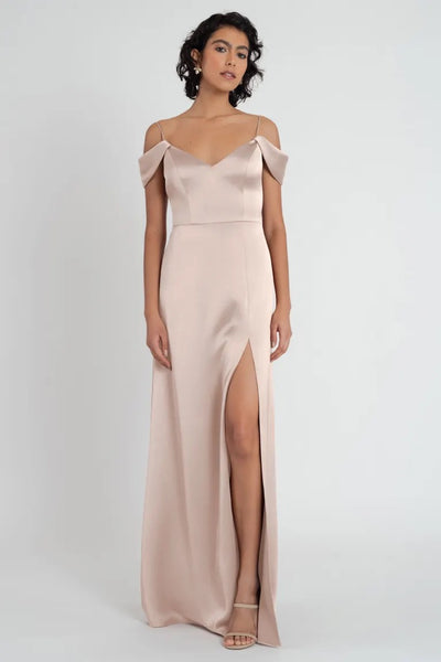 Woman posing in a Priyanka - Bridesmaid Dress by Jenny Yoo from Bergamot Bridal, a blush-toned satin dress with a thigh-high slit and off-the-shoulder sleeves.