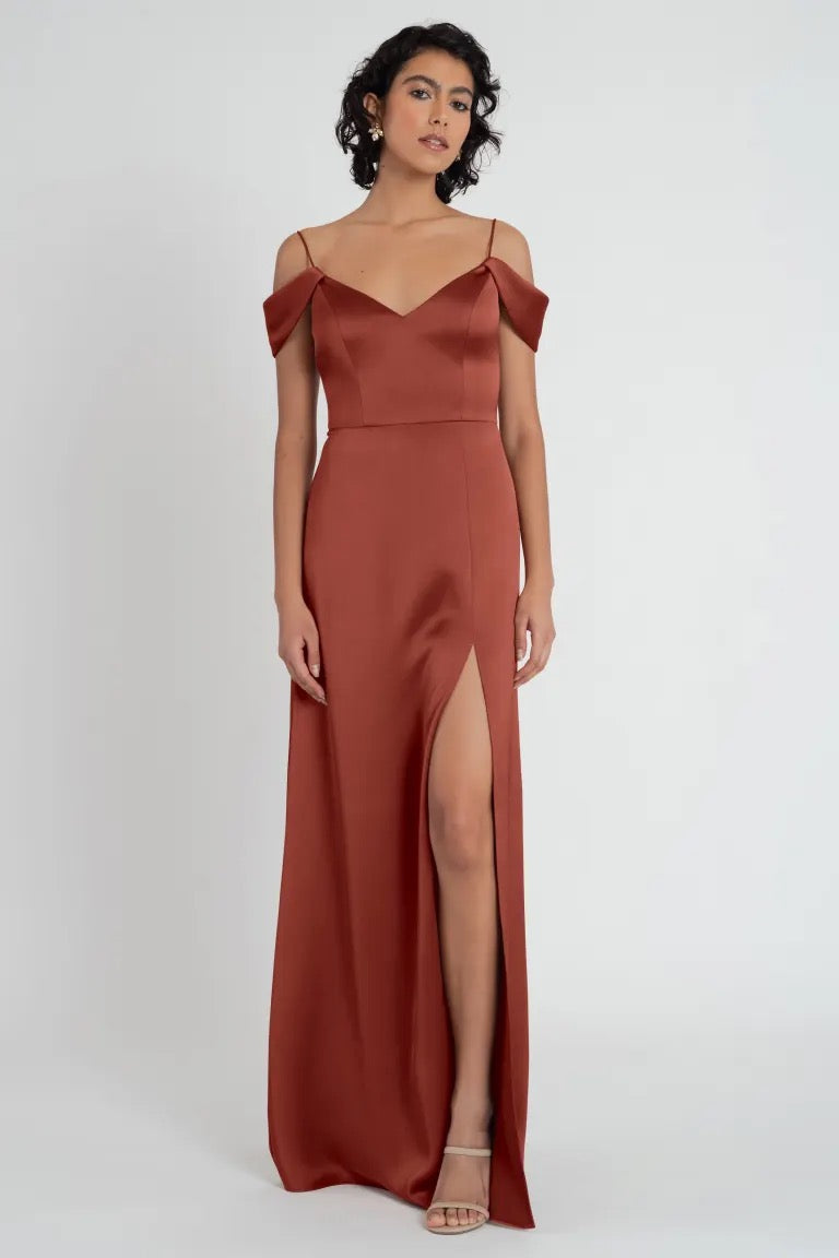 A woman wearing an elegant rust-colored satin Priyanka bridesmaid dress by Jenny Yoo with a high leg slit and off-the-shoulder sleeves from Bergamot Bridal.