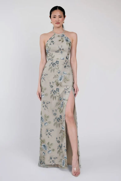 A woman is wearing the Rebecca - Jenny Yoo Bridesmaid Dress by Bergamot Bridal, a floor-length, sleeveless floral dress featuring a high slit and a stylish halter neckline. She stands against a plain background and faces forward.