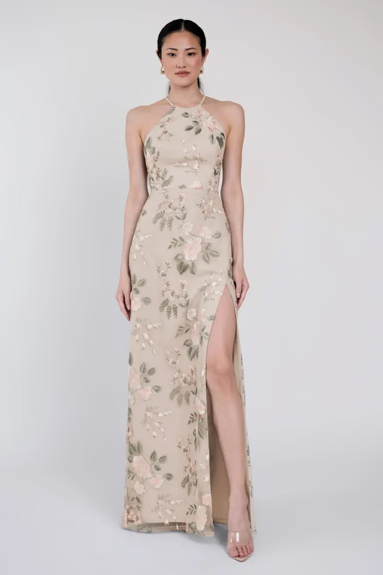 A person stands wearing the Rebecca - Jenny Yoo Bridesmaid Dress by Bergamot Bridal, featuring a sleeveless, high-neck, floral pattern with a thigh-high slit, posing against a plain background.