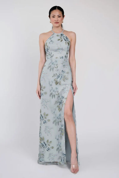 A woman is wearing a light blue Jenny Yoo Bridesmaid Dress by Bergamot Bridal, featuring Ophelia embroidery and a high slit. She is standing against a plain white background.