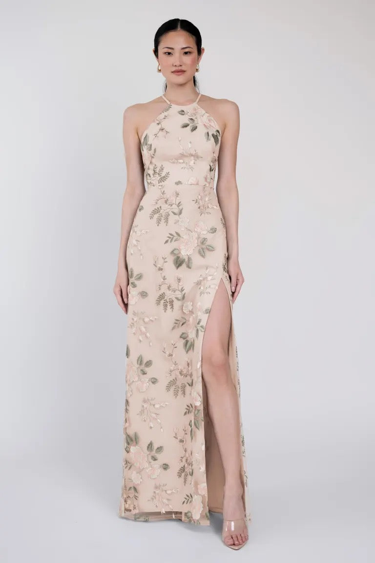 A woman stands wearing a floor-length, sleeveless Jenny Yoo Bridesmaid Dress called "Rebecca" by Bergamot Bridal, featuring a halter neckline and a high slit on the left side. She has her hair tied back and wears clear high heels.