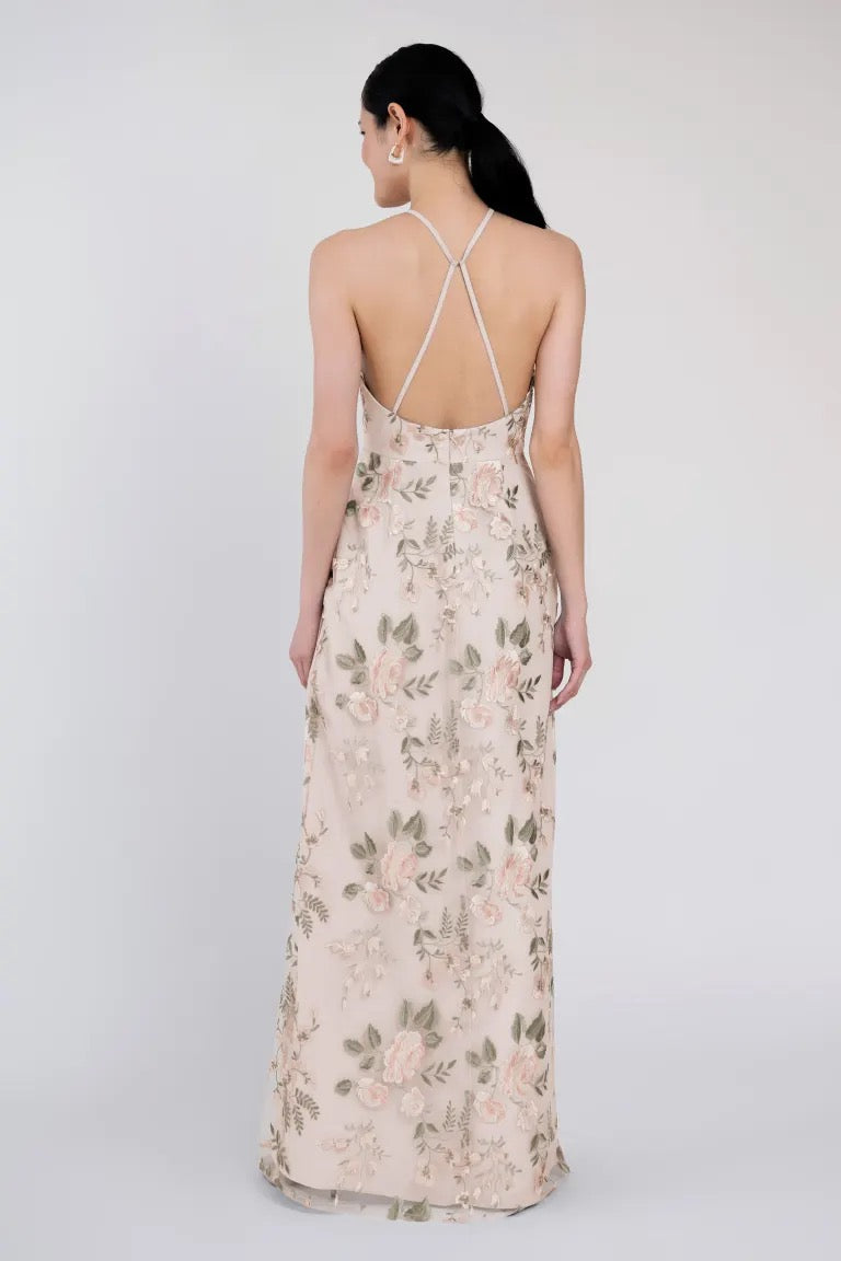 A woman wearing a long, floral Rebecca - Jenny Yoo Bridesmaid Dress by Bergamot Bridal, featuring Ophelia embroidery and a crisscross strap back, is standing facing away from the camera.