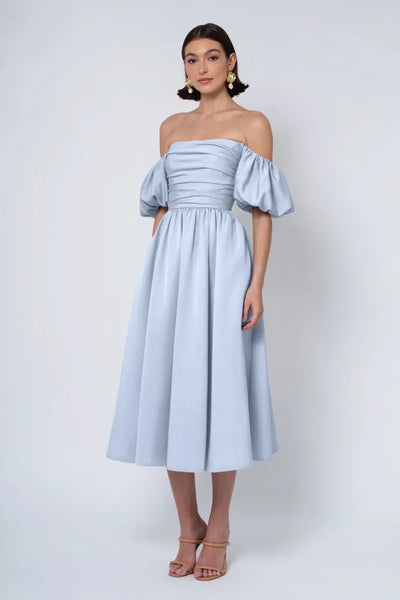 A woman models a pale blue Jenny Yoo Bridesmaid Dress with a strapless neckline and puff sleeves, standing against a white background from Bergamot Bridal.