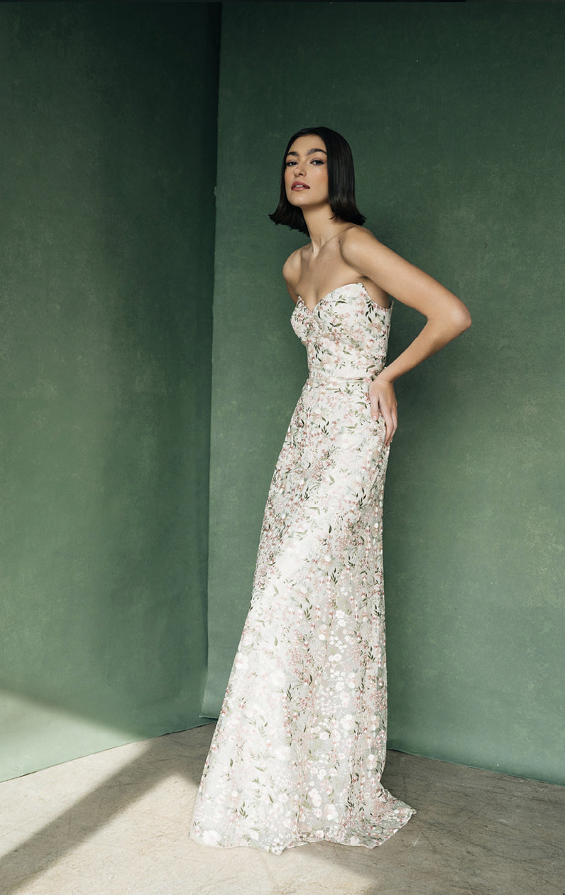 A woman in an elegant Jenny Yoo Bridesmaid Dress with Ophelia embroidery posing against a green wall from Bergamot Bridal.