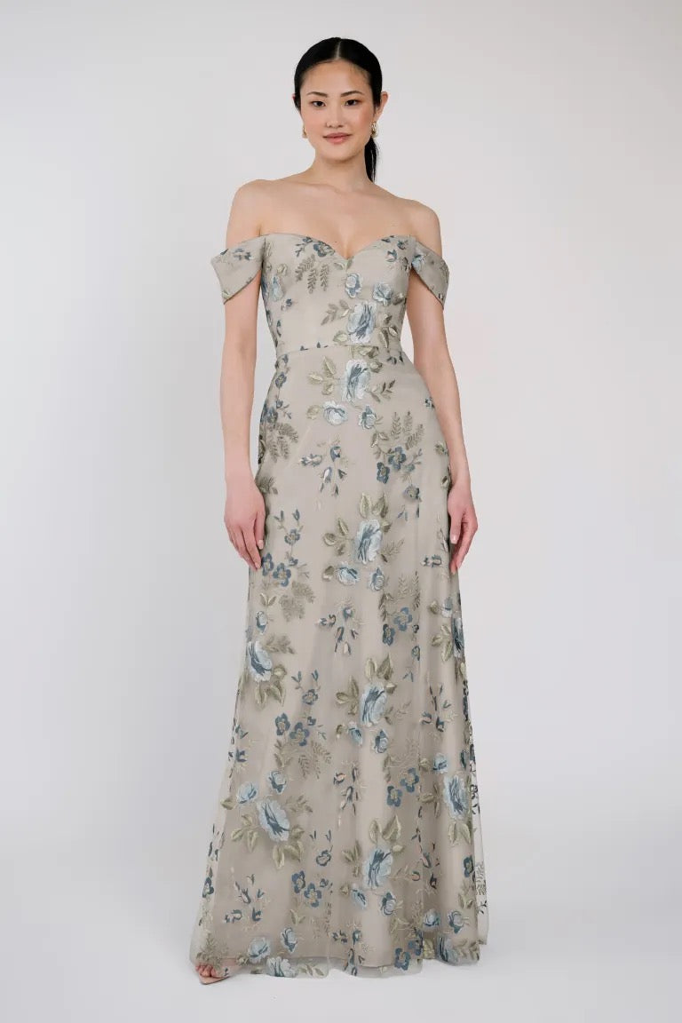 A woman stands wearing an off-shoulder, floor-length Reina - Jenny Yoo Bridesmaid Dress in beige with blue floral patterns, accented by delicate Ophelia embroidery from Bergamot Bridal.