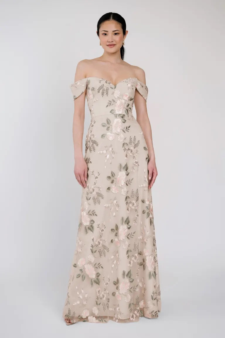 A woman in a strapless, beige, floor-length Reina dress by Jenny Yoo with a sweetheart neckline and adorned with Ophelia embroidery from Bergamot Bridal stands against a plain background.