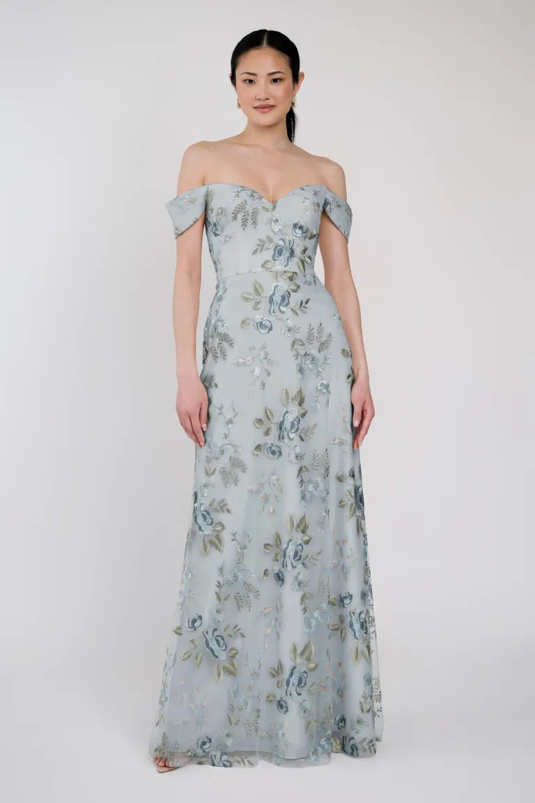 A woman stands wearing the Reina - Jenny Yoo Bridesmaid Dress from Bergamot Bridal, an off-the-shoulder design with floral Ophelia embroidery on a light blue base, featuring a sweetheart neckline, looking forward with a neutral expression.