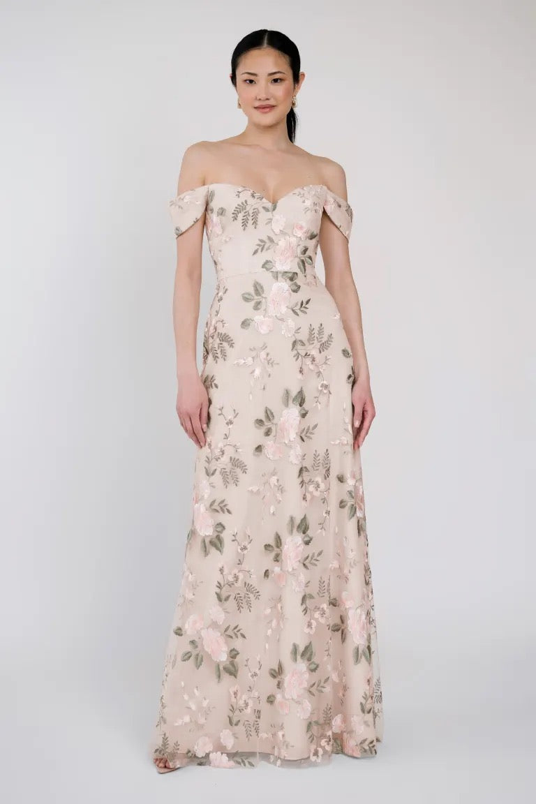 A woman wearing an off-shoulder Reina - Jenny Yoo Bridesmaid Dress from Bergamot Bridal, adorned with Ophelia embroidery and a floral pattern, stands against a plain background.