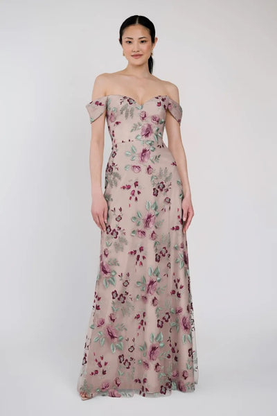 A person wearing an off-the-shoulder, floral-patterned Reina - Jenny Yoo Bridesmaid Dress by Bergamot Bridal stands against a plain, light-colored background.