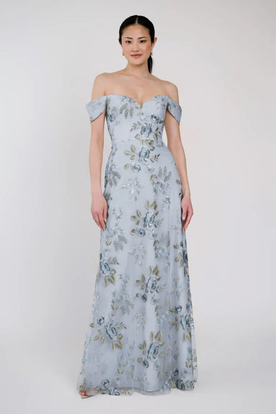 A woman is wearing the Reina - Jenny Yoo Bridesmaid Dress by Bergamot Bridal, a light blue off-the-shoulder gown adorned with Ophelia embroidery and a floral pattern, standing against a plain background.