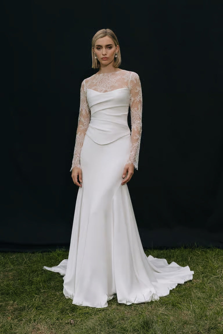 A woman poses on grass before a black backdrop, wearing a long white wedding dress with lace sleeves, a fitted bodice, and the Renata Topper by Jenny Yoo from Bergamot Bridal.