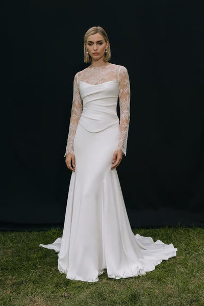 A woman poses on grass before a black backdrop, wearing a long white wedding dress with lace sleeves, a fitted bodice, and the Renata Topper by Jenny Yoo from Bergamot Bridal.