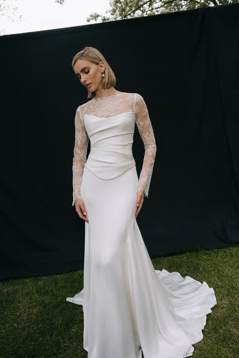 A woman stands on grass in front of a black backdrop wearing a long white wedding dress with lace sleeves and the Renata Topper by Jenny Yoo from Bergamot Bridal.
