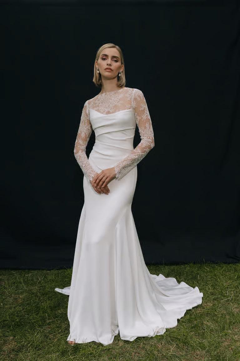 A woman stands on grass in a long white wedding dress with lace sleeves and a fitted bodice, styled with the Renata Topper by Jenny Yoo from Bergamot Bridal, posing against a plain black backdrop.