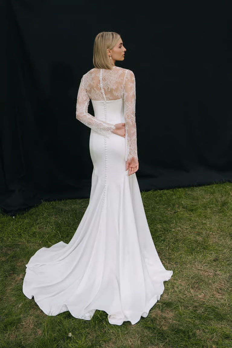 A woman wearing the Renata Topper by Jenny Yoo from Bergamot Bridal stands on grass with her back to the camera, framed by a black backdrop. She pairs a white, long-sleeved lace wedding gown with the delicate bridal topper.