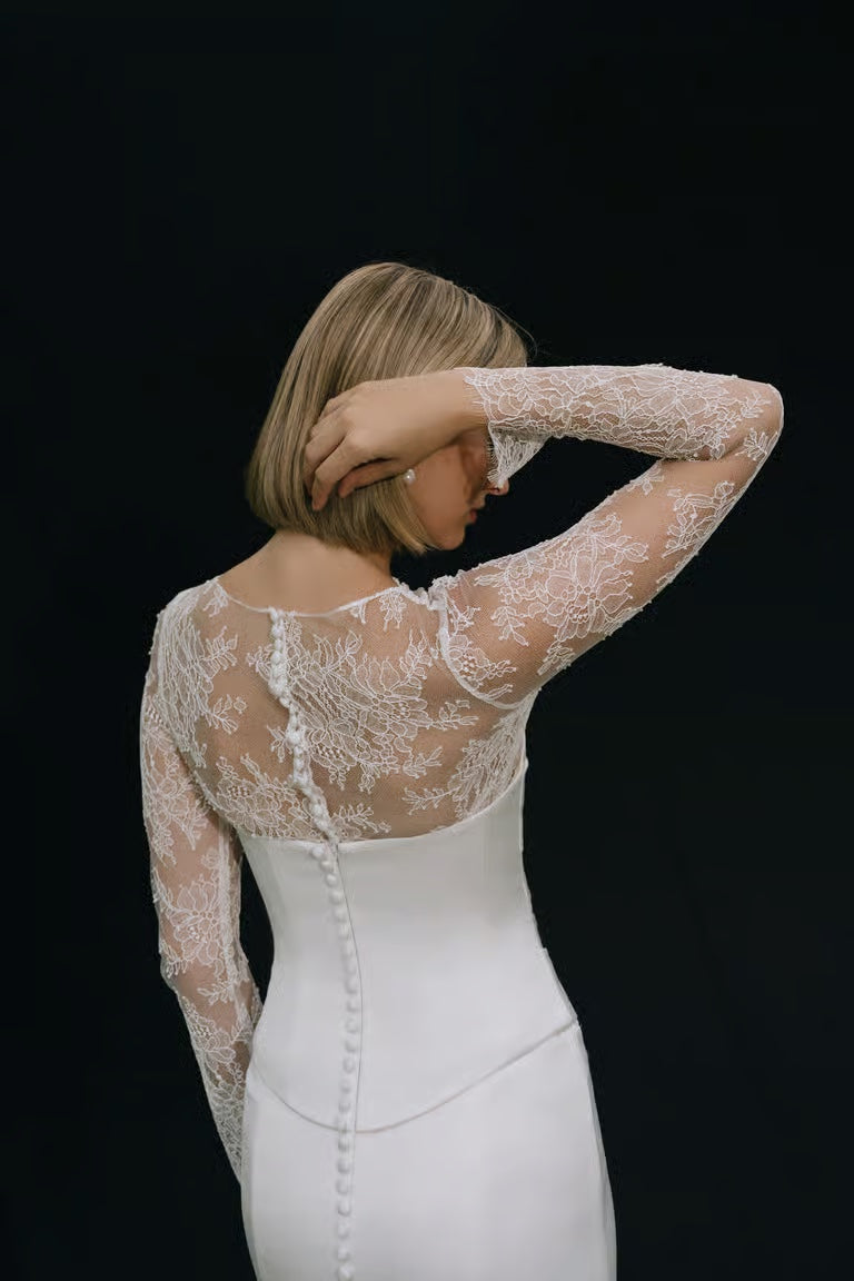 A woman with short blonde hair, seen from behind, wears a white gown with sheer lace sleeves and back detail, elegantly styled with the Renata Topper by Jenny Yoo from Bergamot Bridal as a stunning wedding accessory against a black background.