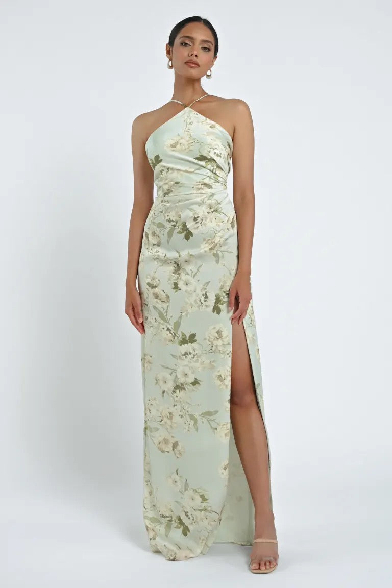 A woman is wearing the Riley Bridesmaid Dress by Jenny Yoo from Bergamot Bridal, a light green floral dress featuring a chic halter neckline and a high slit. She has her hair pulled back and is posing against a plain white background.