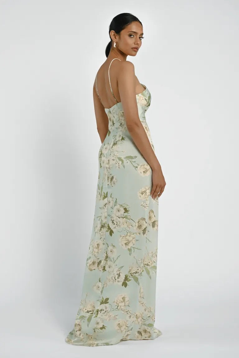 A woman in a light green Riley Bridesmaid Dress by Jenny Yoo from Bergamot Bridal, featuring thin straps and asymmetrical pleats, stands facing away from the camera with her head slightly turned to the side.