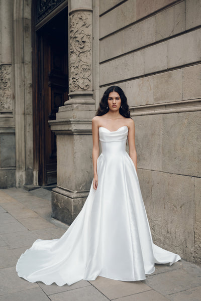 A woman in a Bergamot Bridal Romina - Jenny Yoo Wedding Dress with a strapless design and drop waist silhouette stands outside next to a stone building.