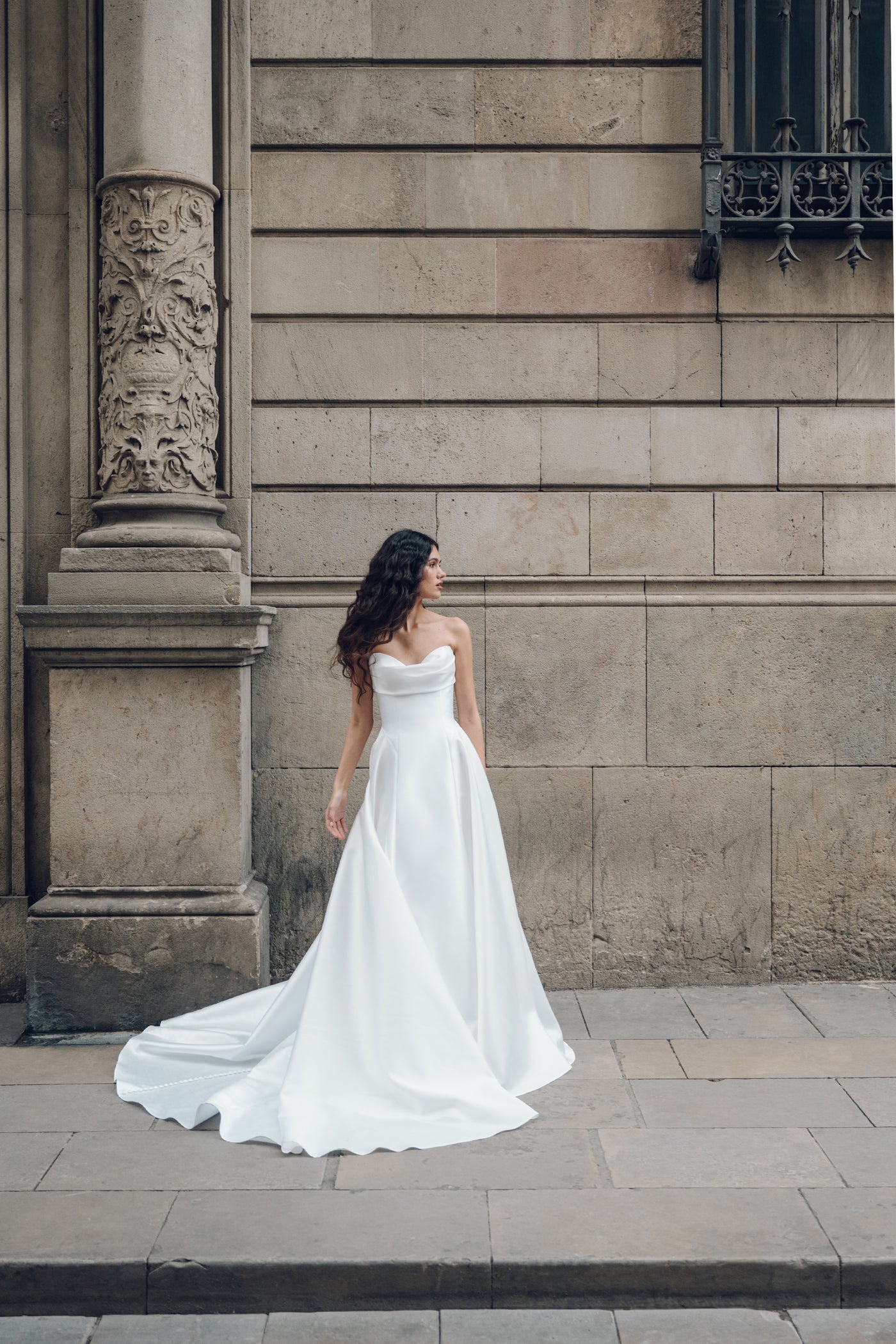 A woman in a strapless Romina - Jenny Yoo Wedding Dress by Bergamot Bridal, featuring a drop waist silhouette, stands on a stone sidewalk next to an ornate column.