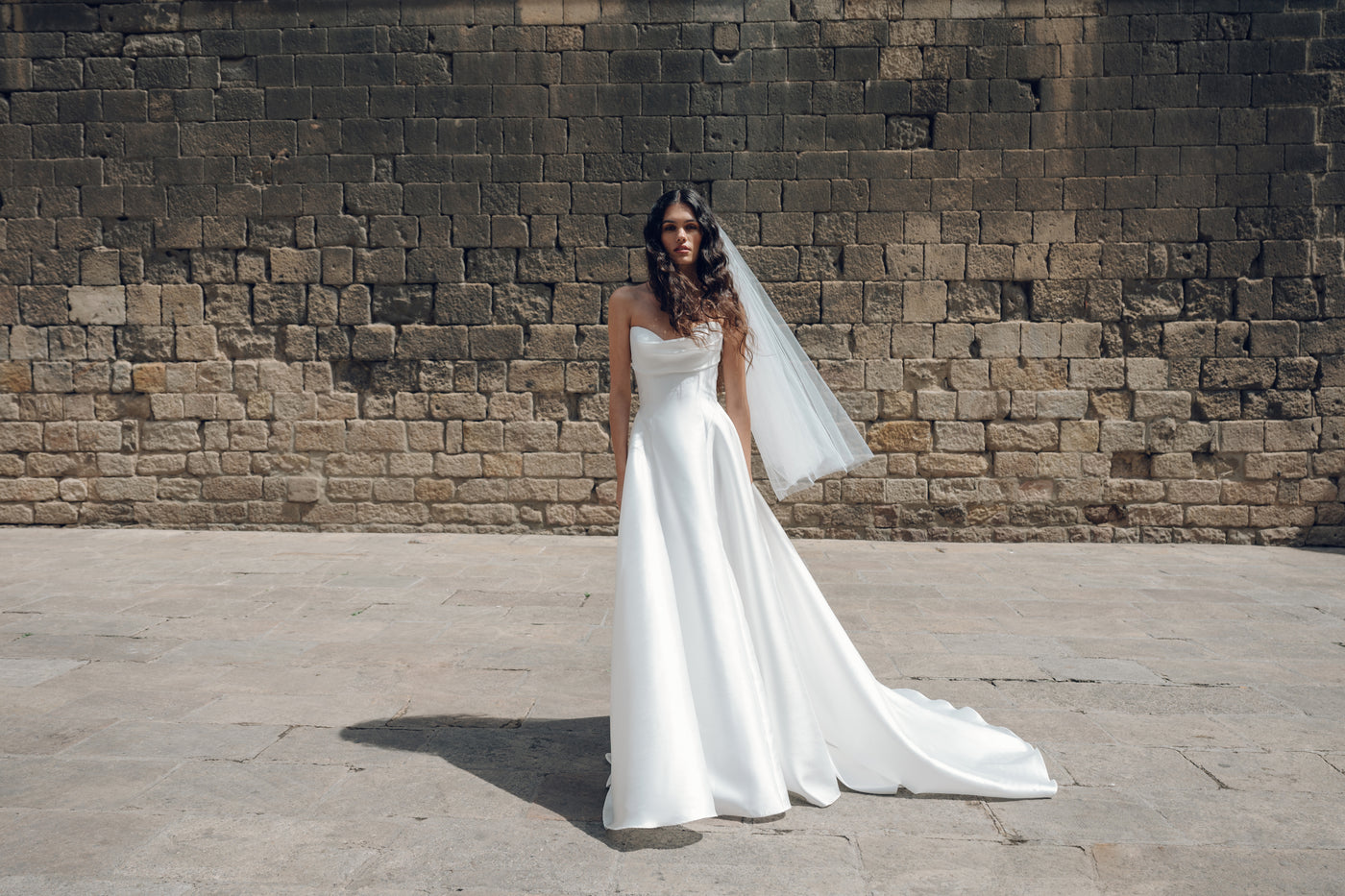 A woman in a strapless Romina gown from Bergamot Bridal, featuring a drop waist silhouette, stands on a stone pavement with her long veil cascading gracefully against a textured stone wall backdrop.