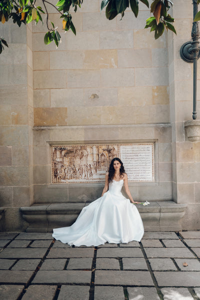 A woman in a Bergamot Bridal Romina – Jenny Yoo Wedding Dress, featuring a taffeta design with a drop waist silhouette, sits against a stone wall adorned with decorative artwork, under the canopy of a tree with green leaves.