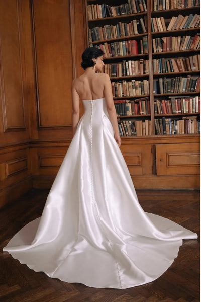 Bride in a white satin Jenny Yoo Wedding Dress with a drop waist silhouette standing in a library from Bergamot Bridal.