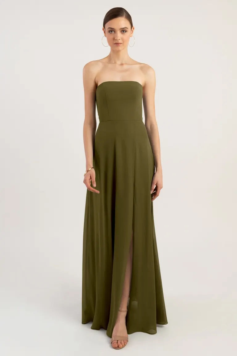 A woman in a strapless chiffon olive green Essie - Bridesmaid Dress by Jenny Yoo with a thigh-high slit stands against a plain background from Bergamot Bridal.