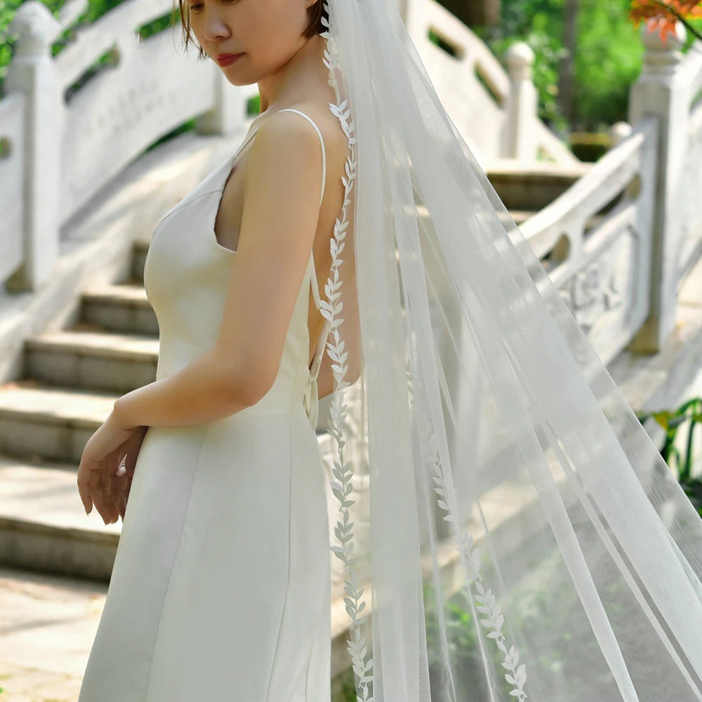 A bride in a white dress, wearing Bergamot Bridal's Leaf Lace Edged Bridal Veil, stands near stone steps and a decorative railing.