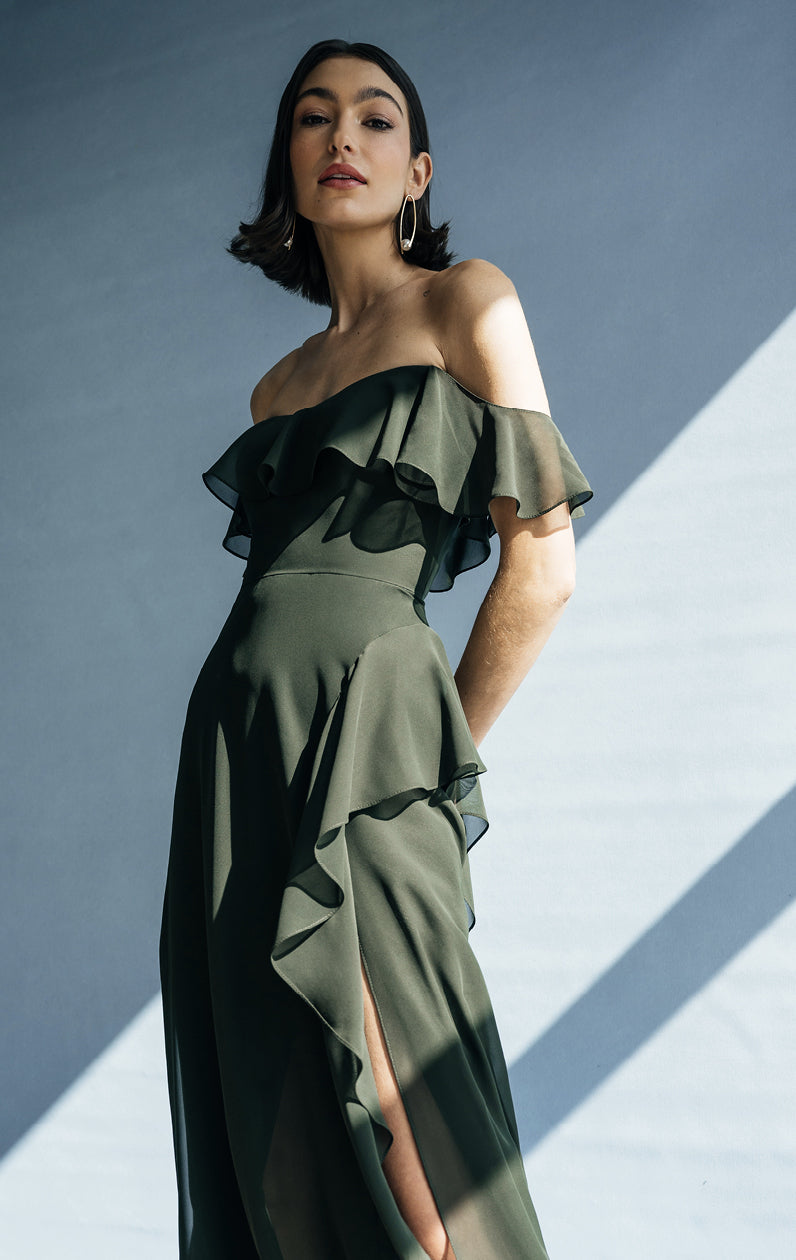 A woman in an elegant Jenny Yoo Bridesmaid Dress from Bergamot Bridal with an off-the-shoulder neckline and ruffle details stands against a background with geometric shadows.