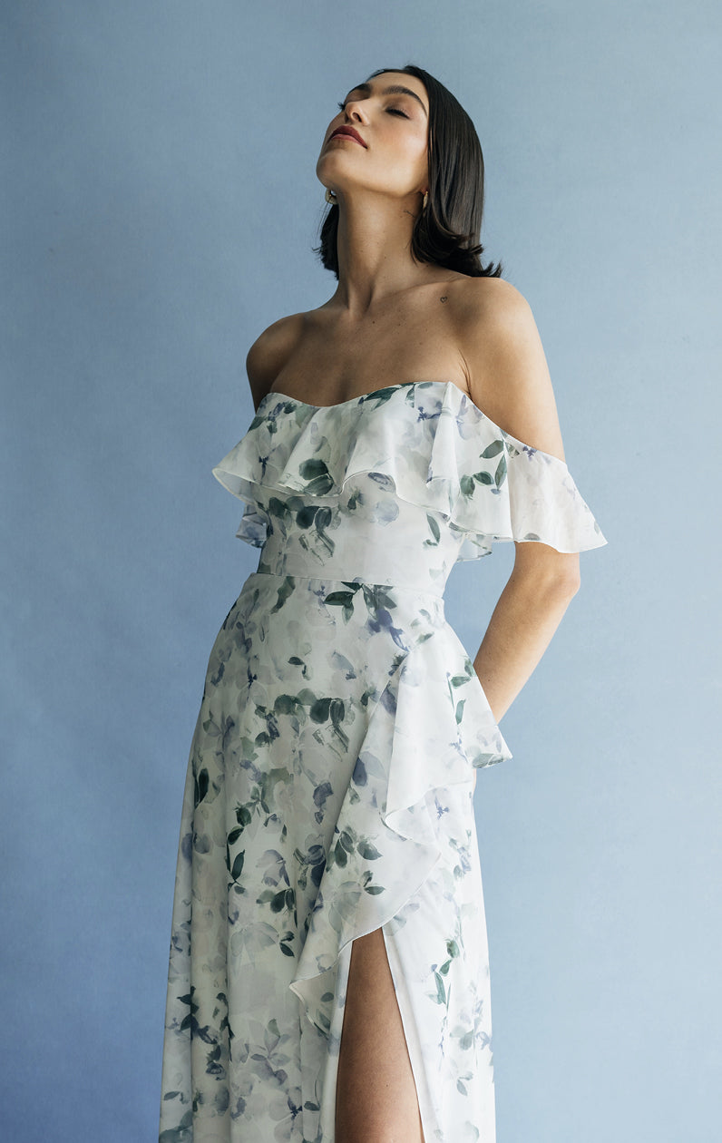 A woman in a Salma Print - Jenny Yoo Bridesmaid Dress by Bergamot Bridal, featuring an off-the-shoulder neckline and printed chiffon fabric, standing against a blue background with her head tilted upwards.