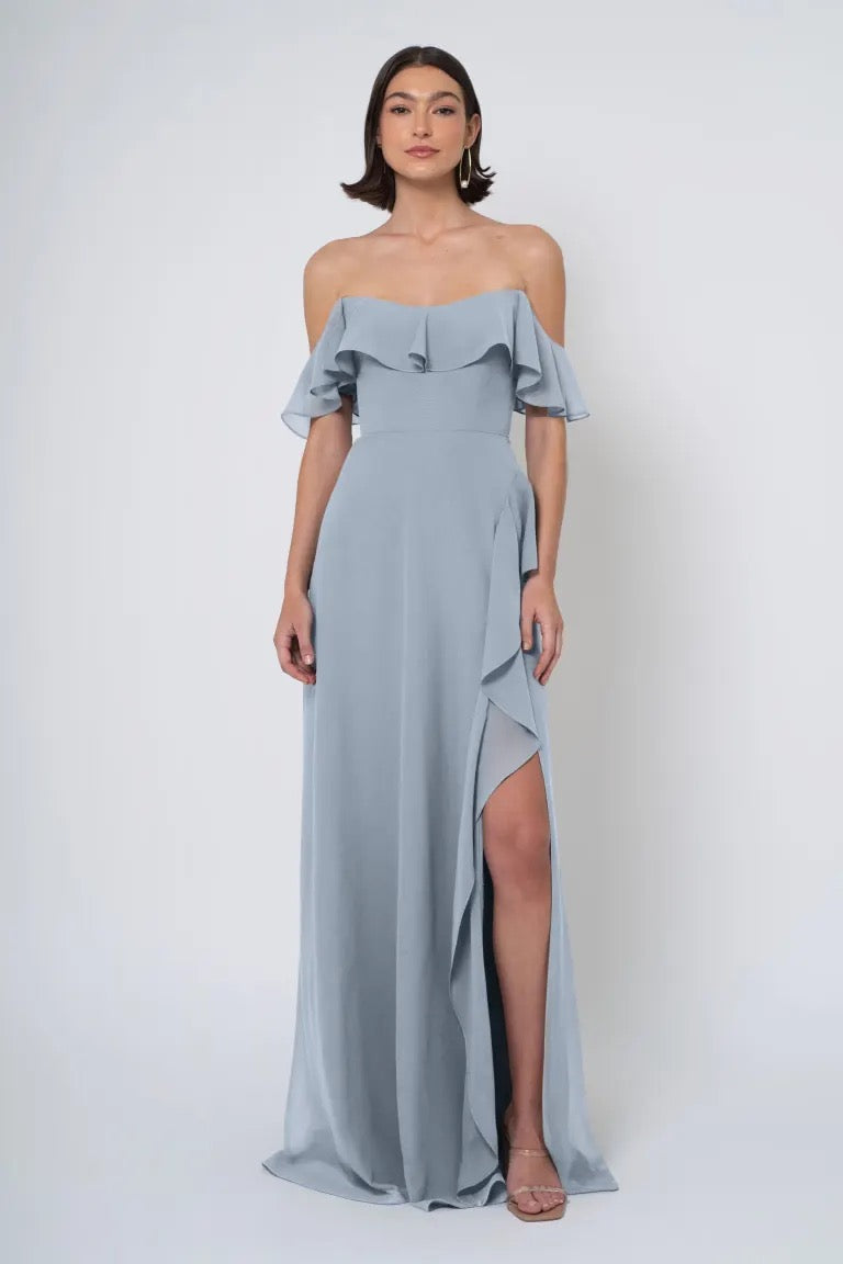 A woman wearing a grey off-the-shoulder neckline Salma chiffon dress with a side slit by Bergamot Bridal.