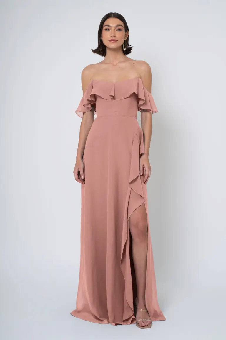 Woman posing in an elegant Salma chiffon dress by Jenny Yoo Bridesmaid Dress with a dusty pink off the shoulder neckline, ruffled sleeves, and a high side slit from Bergamot Bridal.