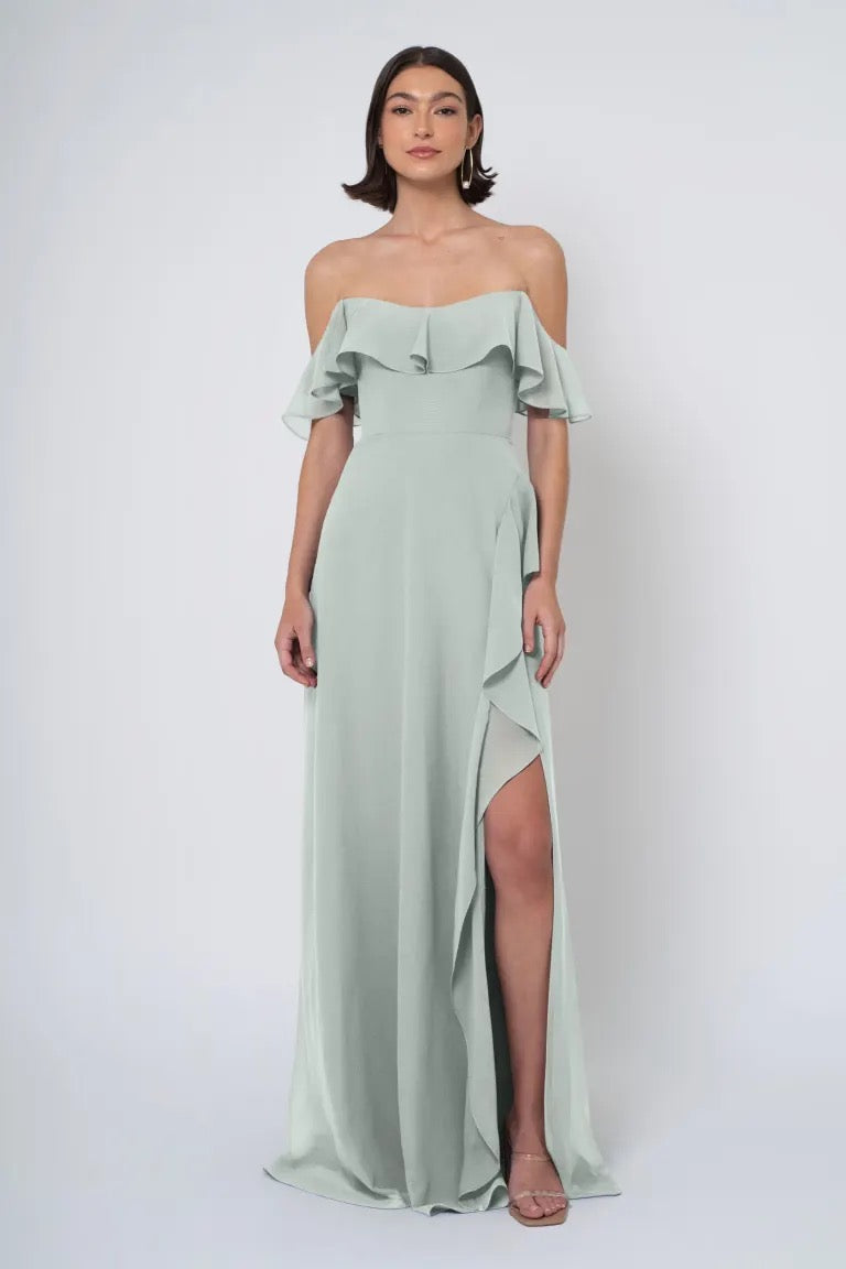 Woman in a sage green Salma - Jenny Yoo Bridesmaid dress with an off-the-shoulder neckline and a thigh-high side slit from Bergamot Bridal.