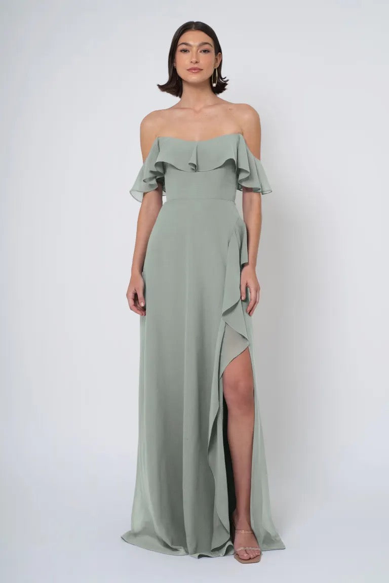 A woman wearing an elegant, off-the-shoulder green Salma - Jenny Yoo Bridesmaid Dress chiffon dress with ruffle details and a side slit from Bergamot Bridal.