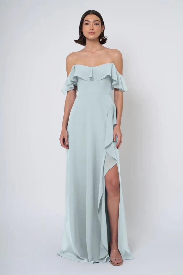 A woman in a light blue Jenny Yoo Bridesmaid Dress, featuring an off-the-shoulder neckline with a side slit, stands against a white background.