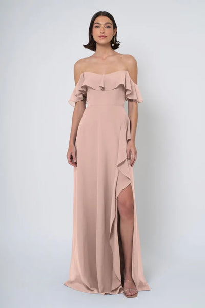 A woman in an elegant, blush-toned Jenny Yoo Bridesmaid Dress with a ruffled off-the-shoulder neckline and a high side slit from Bergamot Bridal.