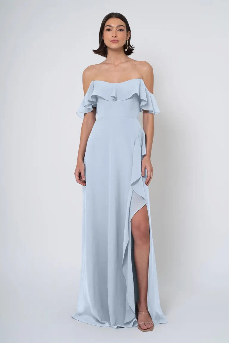 A woman in an elegant, light blue Salma - Jenny Yoo Bridesmaid Dress with a ruffled neckline and a side slit from Bergamot Bridal.