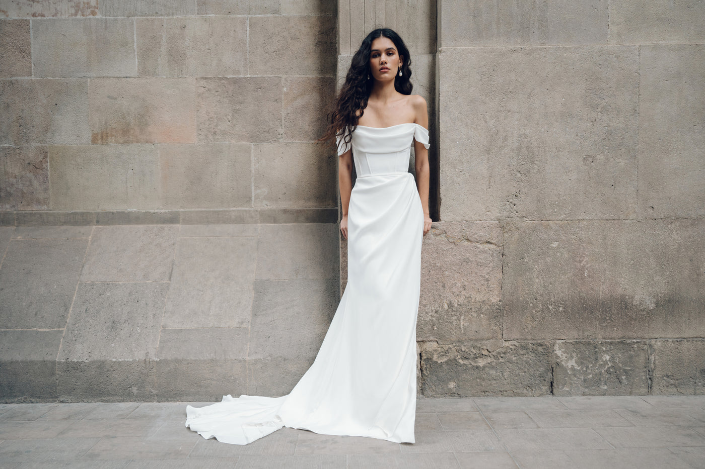 A woman in an off-shoulder white dress with draped details stands against a stone wall, the skirt of her Sandra gown from Bergamot Bridal gracefully trailing on the ground.