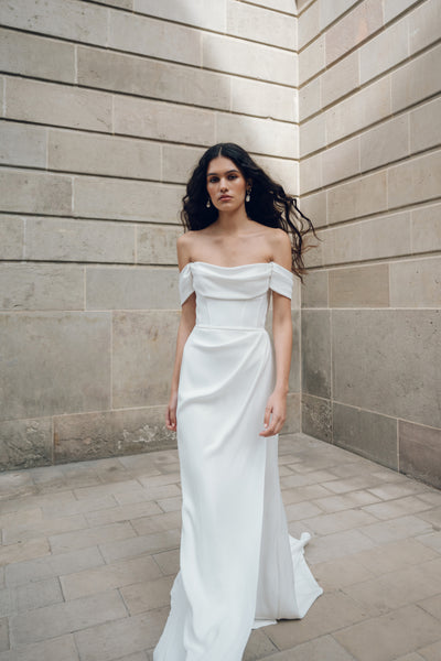 A woman in the Sandra - Jenny Yoo Wedding Dress from Bergamot Bridal, reminiscent of a Greek statue's elegance, stands against a stone wall. Delicate corset details gracefully accentuate her silhouette, adding charm and sophistication to the scene.