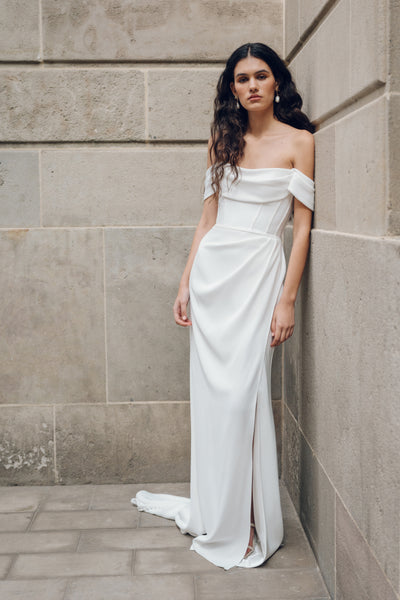 Dressed in a Sandra gown by Jenny Yoo, part of the Bergamot Bridal collection, a woman leans gracefully against a stone wall. The off-the-shoulder white dress with its corset details gives her the appearance of a Greek statue as she stands on the tiled floor.