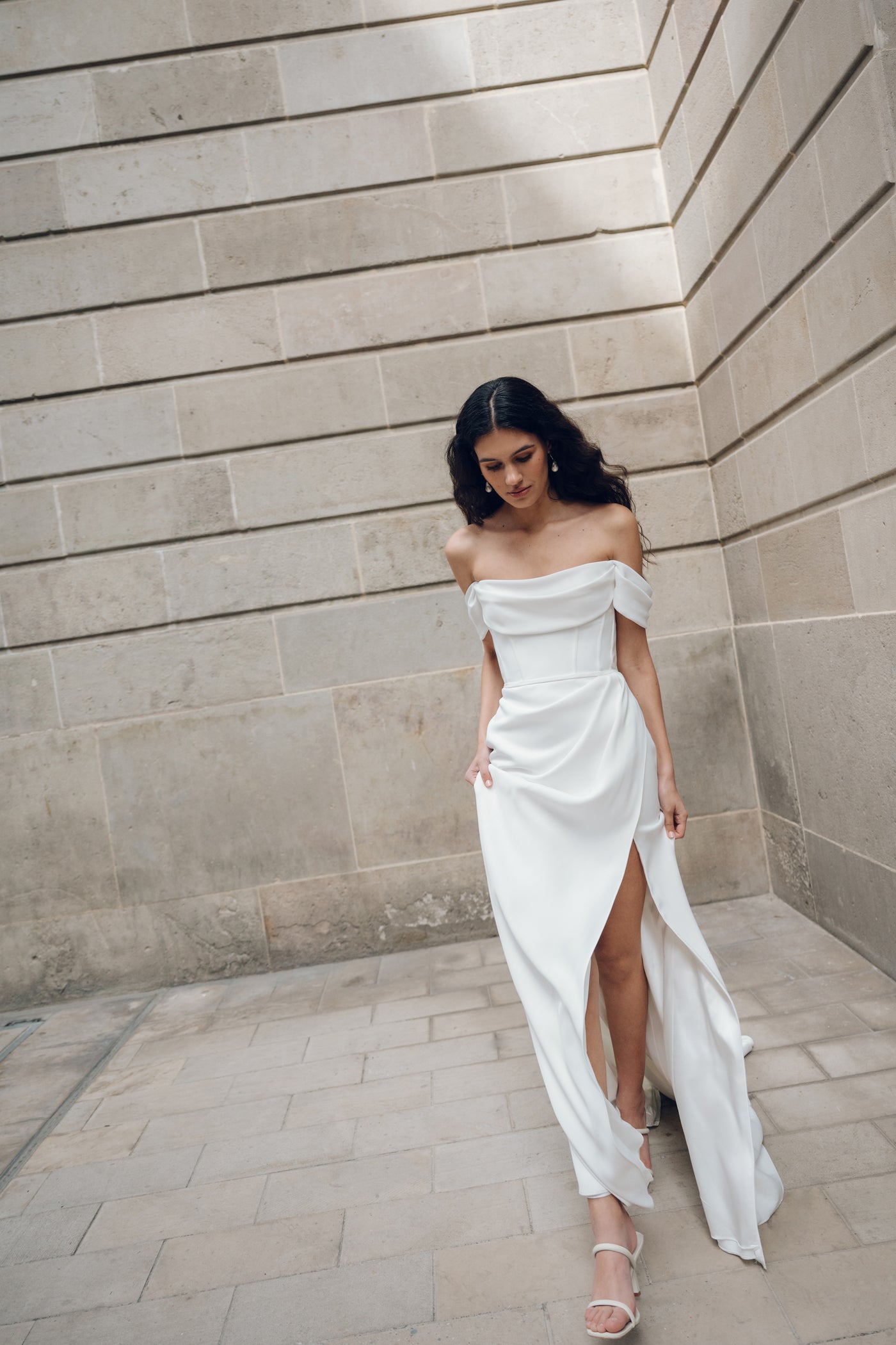 A woman in an off-the-shoulder white Jenny Yoo Wedding Dress named Sandra, featuring corset details and a thigh-high slit, gracefully walks beside a stone wall. This elegant gown is from the Bergamot Bridal collection.