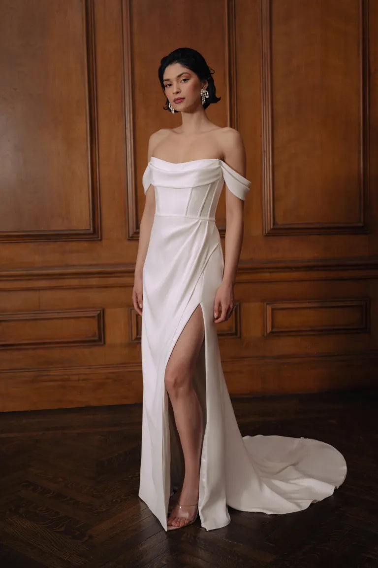 A woman in an elegant off-shoulder Sandra - Jenny Yoo Wedding Dress gown with corset details and a high slit stands against a wooden paneled background from Bergamot Bridal.