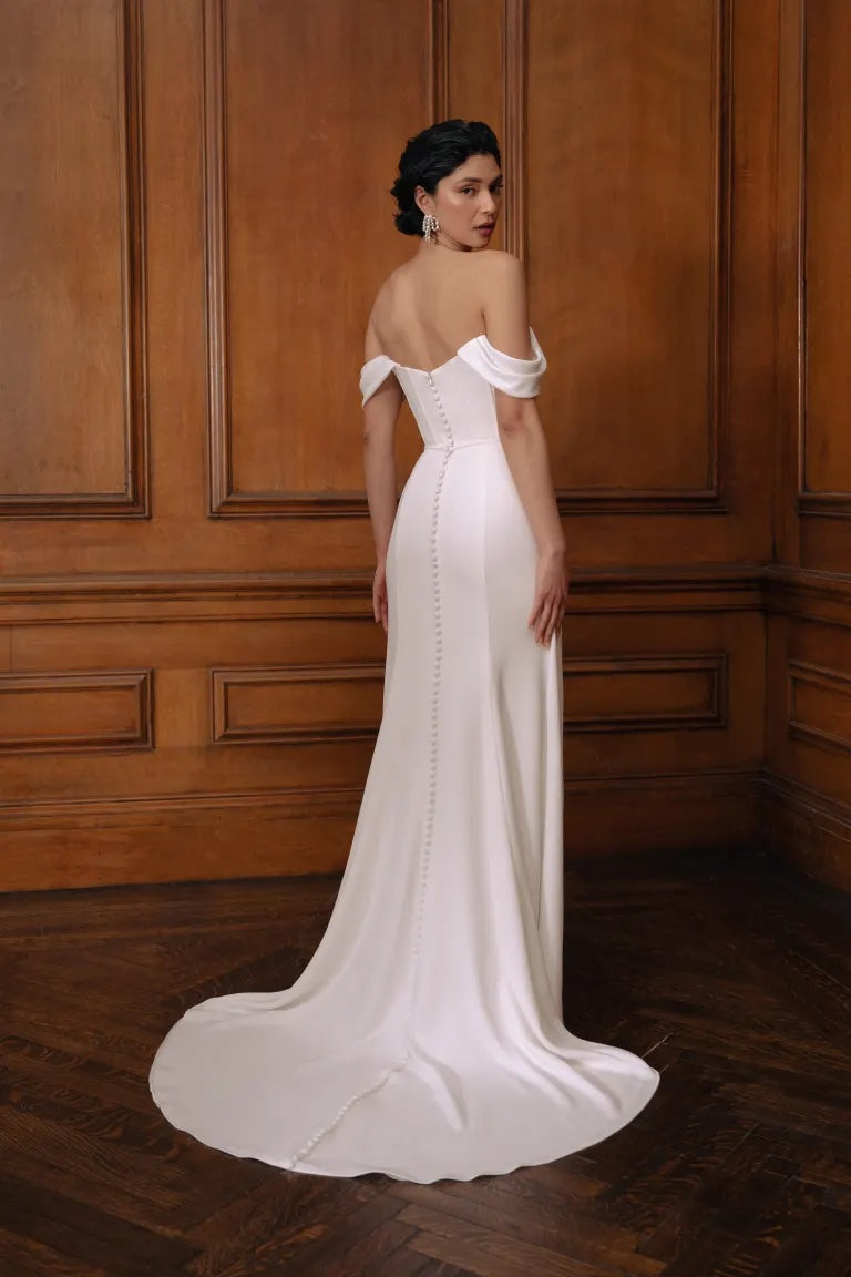 A woman stands before wood-paneled walls in Sandra by Jenny Yoo—an off-the-shoulder wedding gown with a pleated bodice, buttoned back, and flowing train from Bergamot Bridal—elegantly looking over her shoulder.