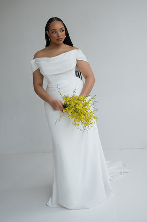 A woman in the Sandra - Jenny Yoo Wedding Dress by Bergamot Bridal, featuring a pleated bodice and sultry slit, holds a yellow bouquet against a plain white background.