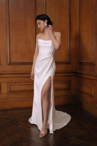 Woman posing in a Jenny Yoo Wedding Dress gown, featuring a pleated bodice and a thigh-high slit, standing against a wooden panel background.