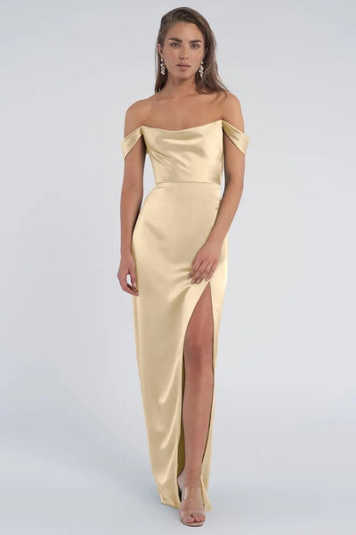 Woman posing in an elegant cowl neckline, off-the-shoulder Sawyer bridesmaid dress by Jenny Yoo in gold luxe satin from Bergamot Bridal with a thigh-high slit.