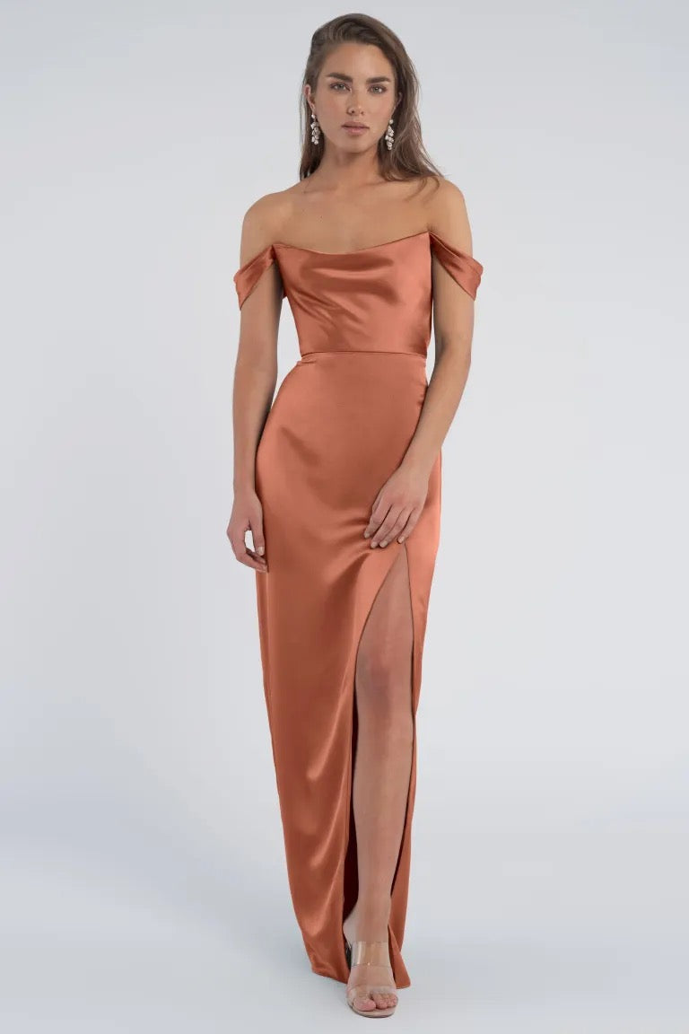 Woman standing in an off-the-shoulder sleeves, luxe satin evening gown with a slit, the Sawyer Bridesmaid Dress by Jenny Yoo from Bergamot Bridal.