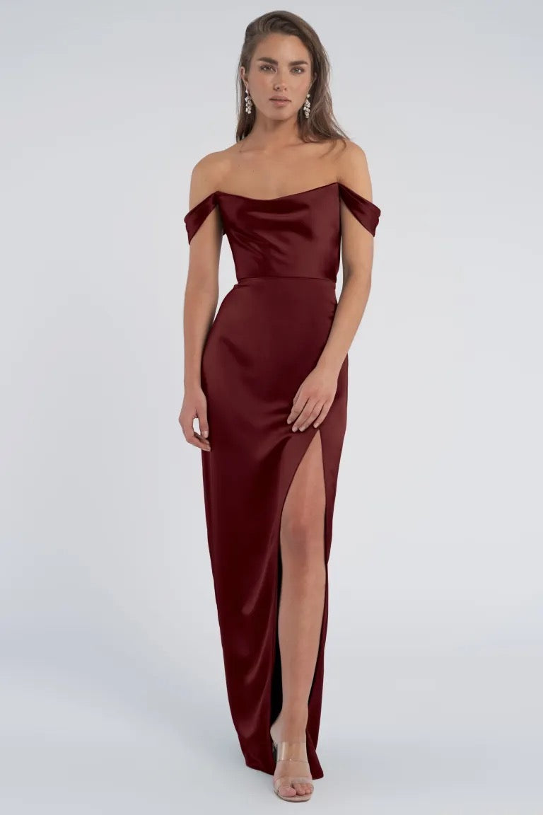 A woman posing in an elegant burgundy off-the-shoulder luxe satin dress with a thigh-high cutaway slit - the Sawyer Bridesmaid Dress by Jenny Yoo from Bergamot Bridal.